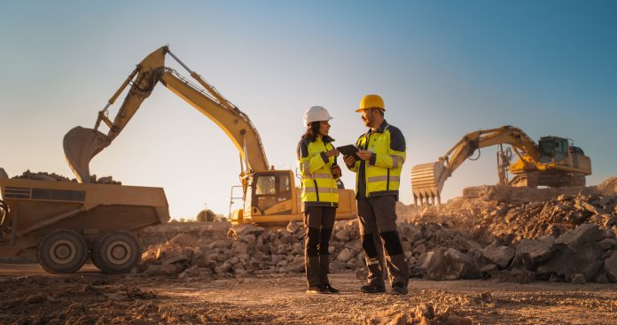 Hispanic Female Inspector Talking to Caucasian Male Land Development Manager With Tablet On Construction Site Of Real Estate Project. Excavators Preparing For Laying Foundation. Hot Sunny Day filière construction